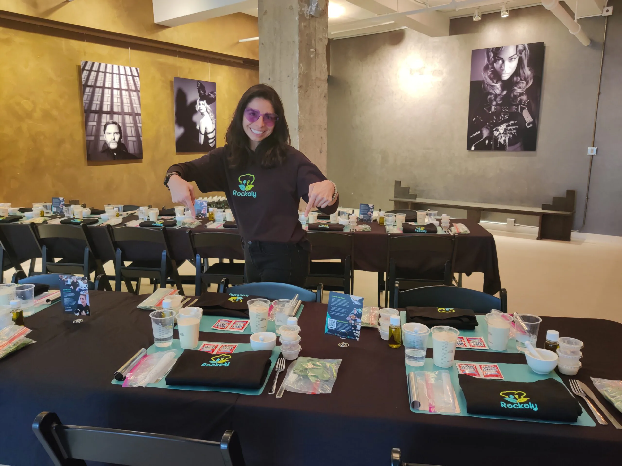 Smiling event host stands with arms open at long tables set with cups, aprons and place mats