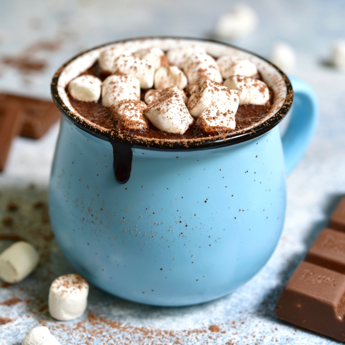 Blue enamel mug of hot chocolate topped with marshmallows and cocoa powder beside chocolate bars
