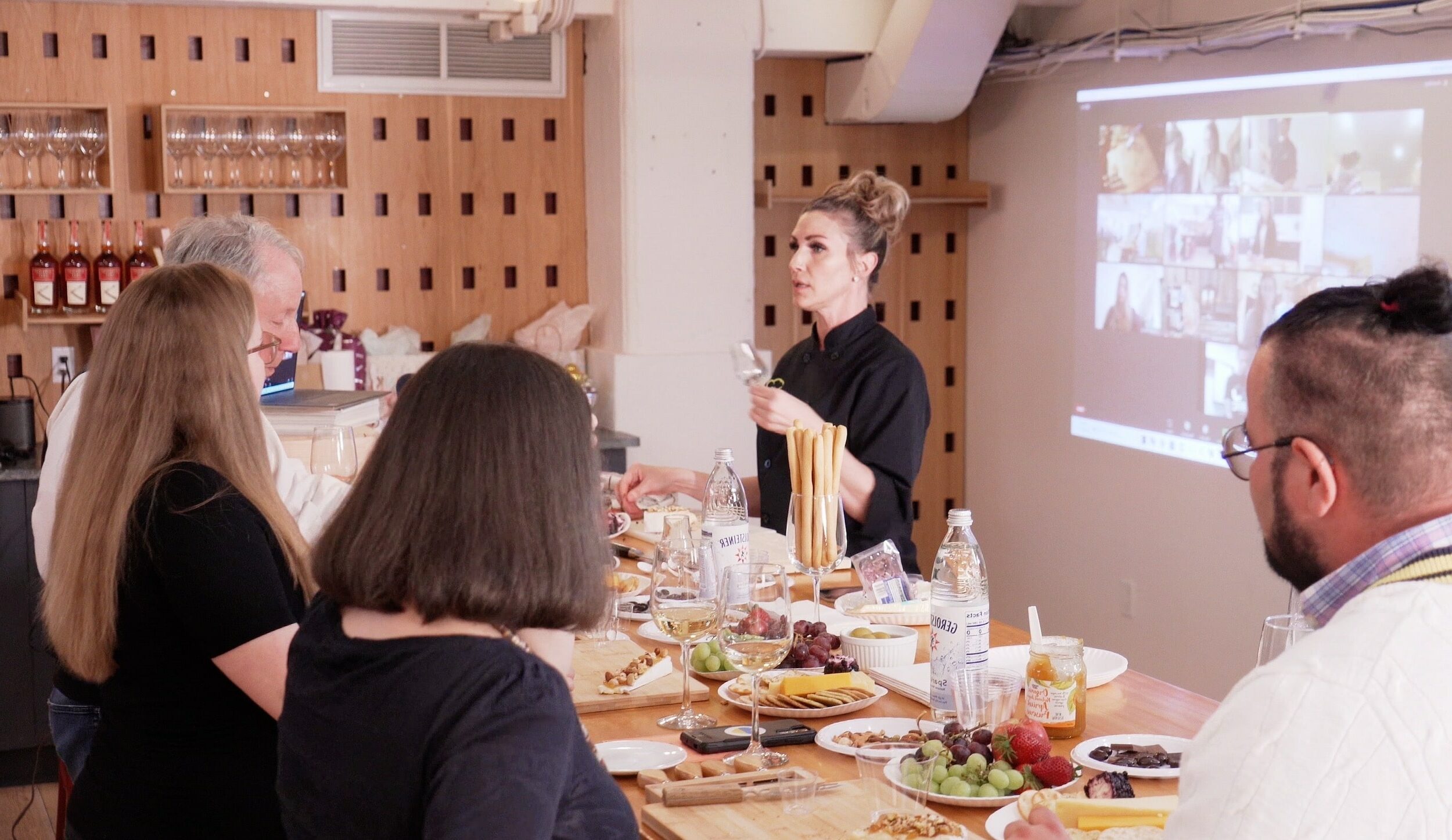 Chef demonstrating fresh pasta to guests at a long table while remote colleagues watch on a projected call