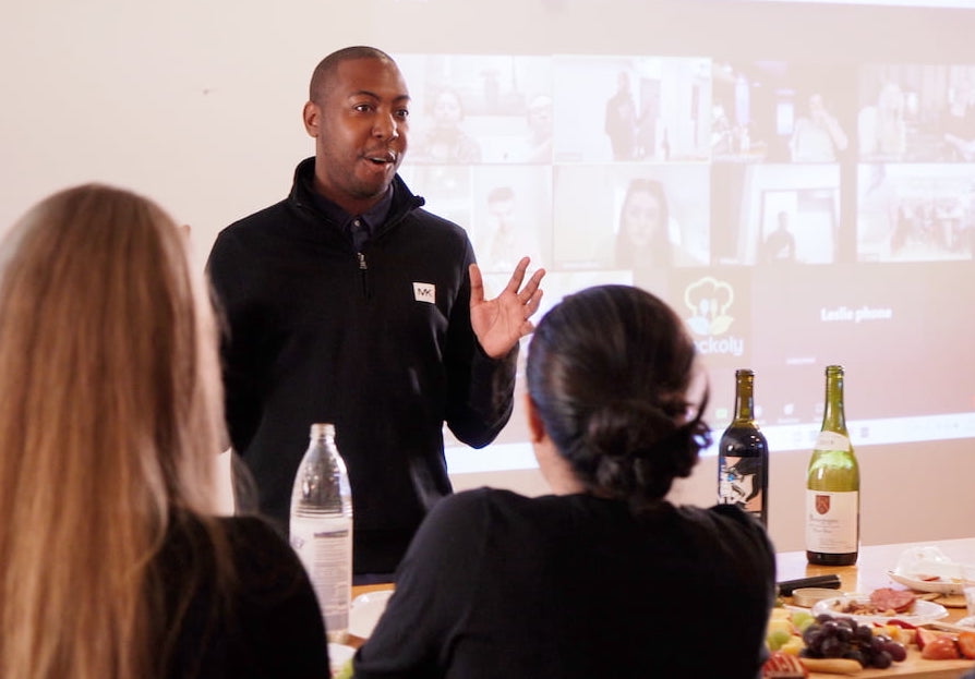 Host presenting to seated guests in front of a projected video call grid, with wine and charcuterie