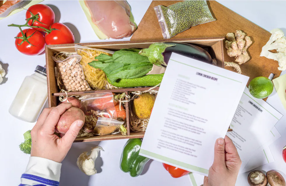 Hands holding a recipe card over an open ingredient kit box of fresh vegetables, chicken and spices
