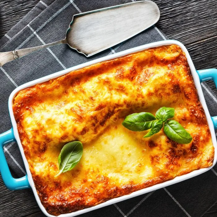 A baked lasagna browned and bubbling in a turquoise ceramic dish, topped with fresh basil