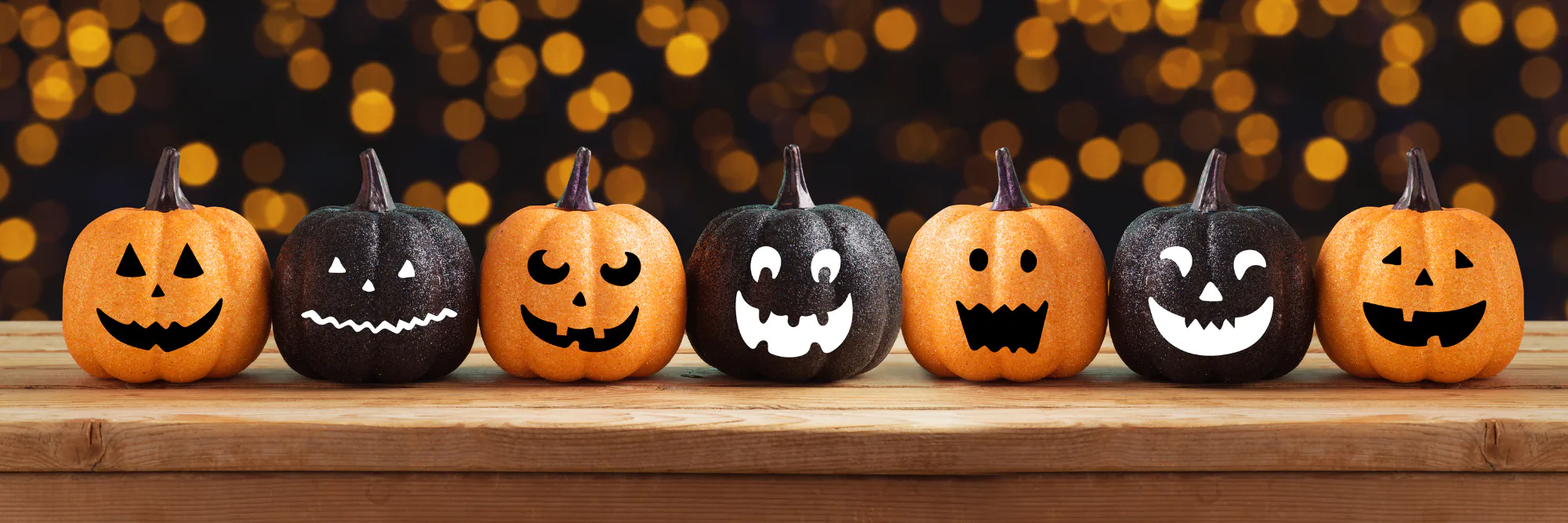 Row of orange and black jack-o'-lanterns on a wooden shelf against warm bokeh lights