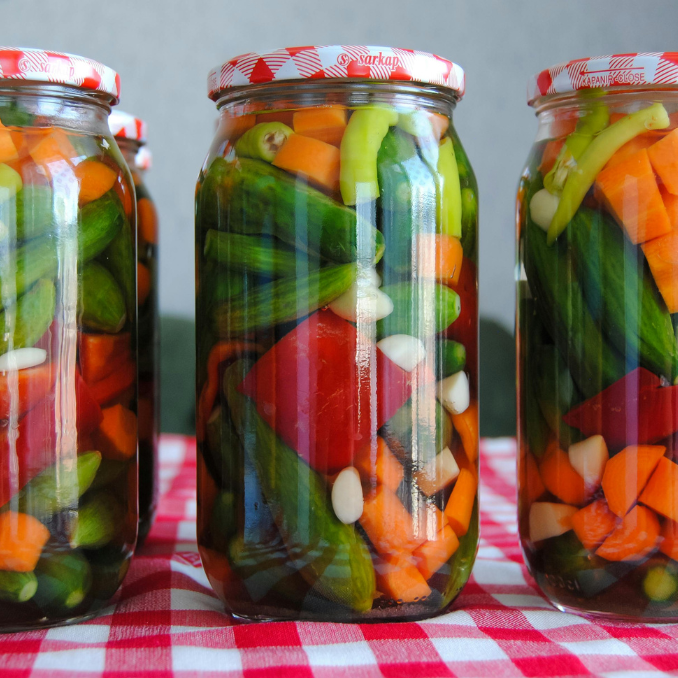 Three glass jars packed with pickled peppers, cucumbers and carrots on a red gingham cloth