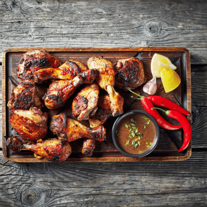 Charred jerk chicken drumsticks and thighs on a wooden board with a bowl of dipping sauce and chilies