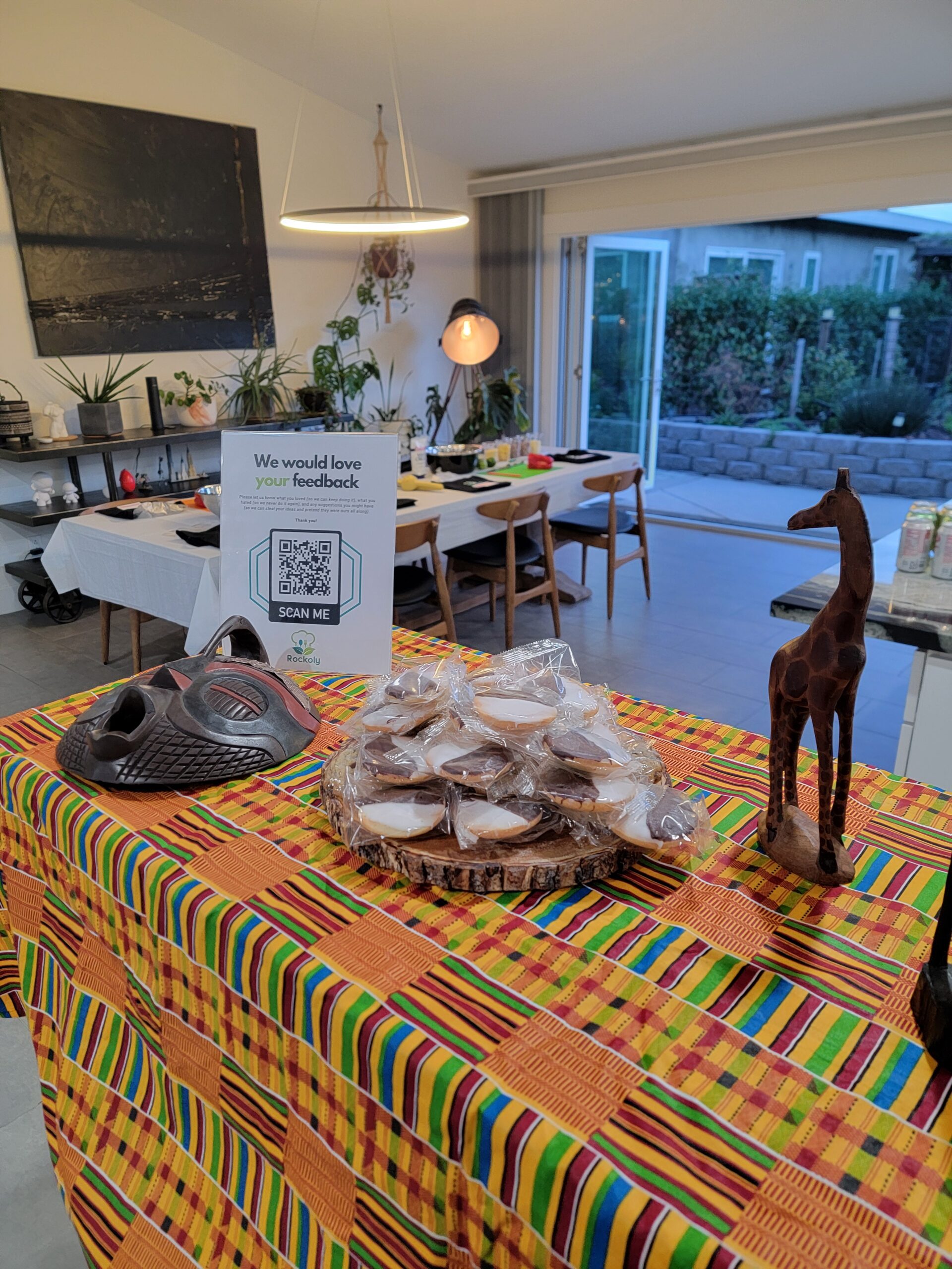 Kente-cloth table holding a tray of wrapped fritters, a carved mask, a wooden giraffe and a feedback sign
