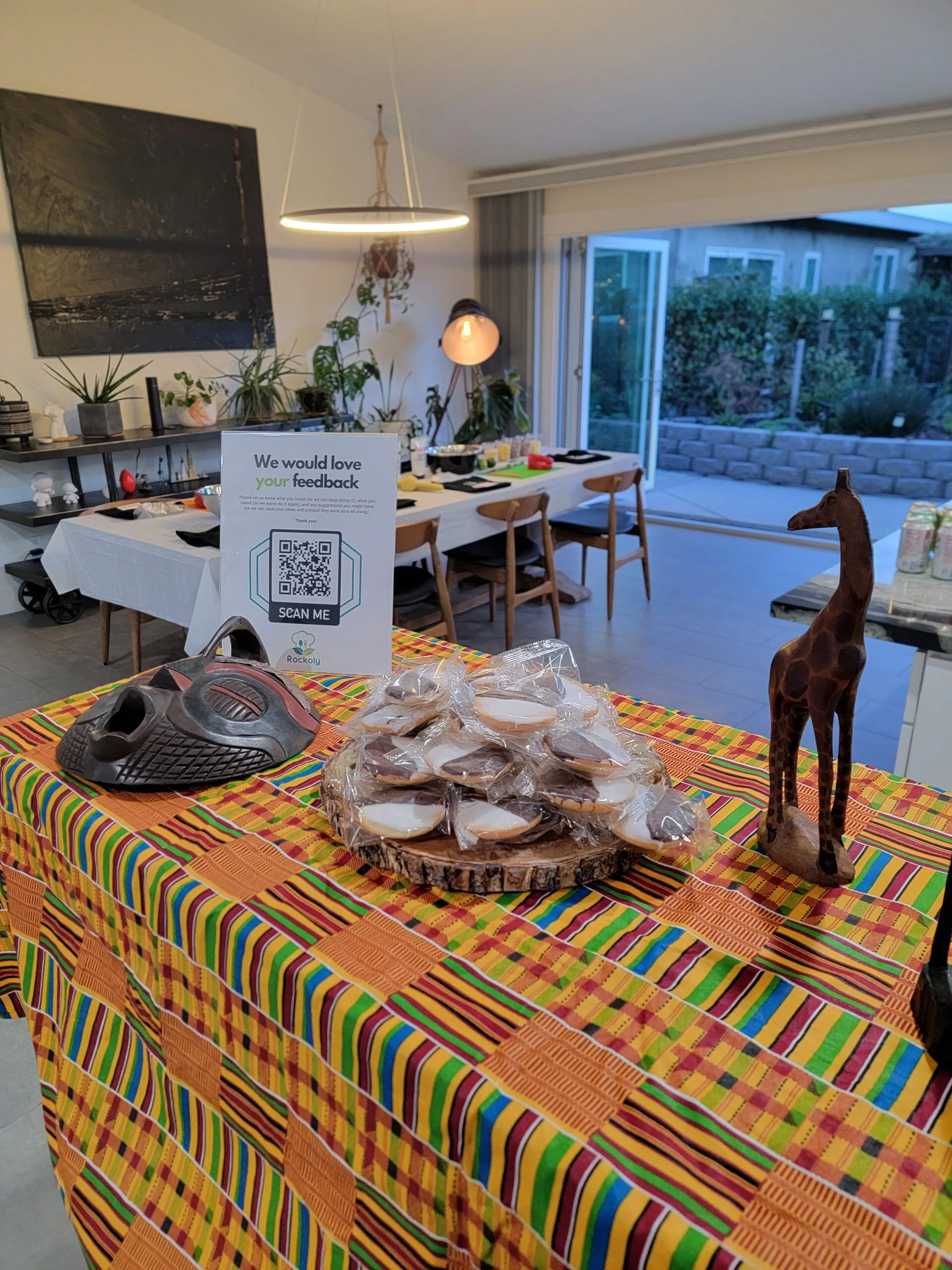Kente-cloth table holding a tray of wrapped fritters, a carved mask, a wooden giraffe and a feedback sign