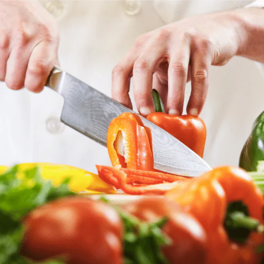 Hands slicing an orange bell pepper with a chef's knife on a board of fresh vegetables