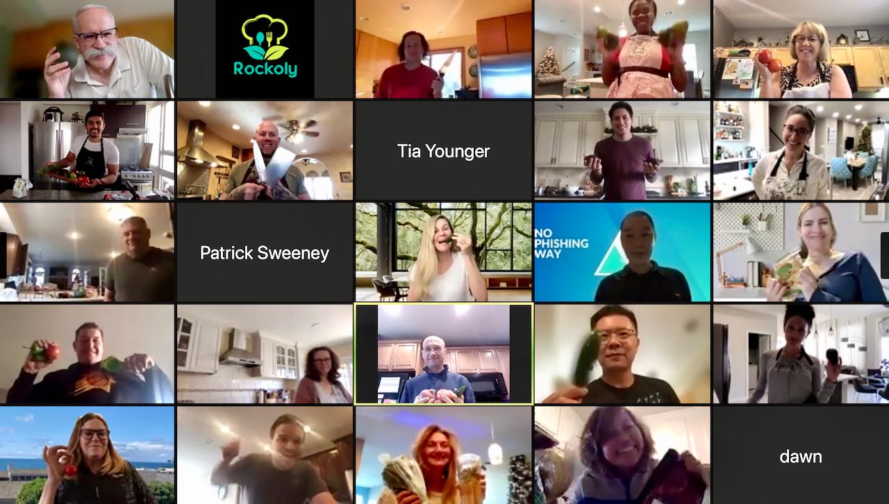 Grid of about twenty coworkers on a video-call cooking class, several raising glasses and utensils