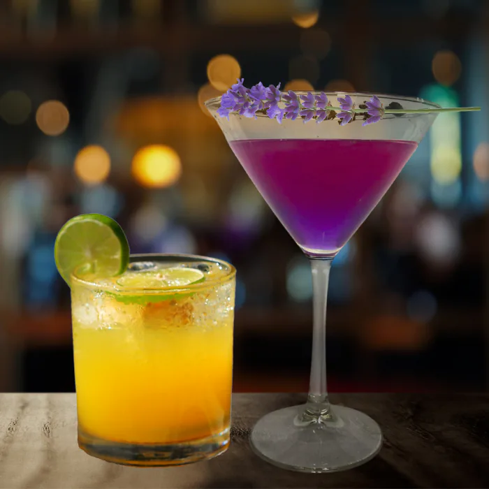 Purple martini topped with lavender sprigs beside an orange cocktail with a lime wheel on a bar