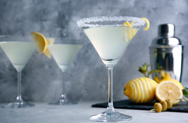 Lemon drop martini with a sugared rim and lemon twist, with a shaker and fresh lemons behind