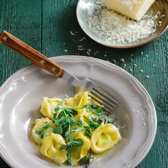 Plate of tortellini tossed with fresh green herbs and grated Parmesan beside a wedge of cheese