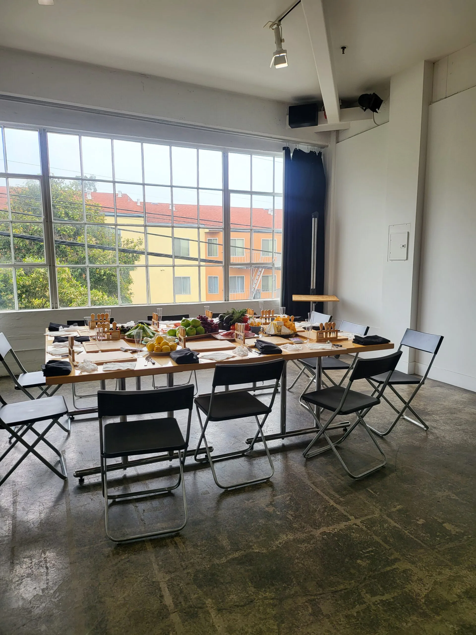 Long table set with ingredients and folding chairs beside tall windows before a cooking event