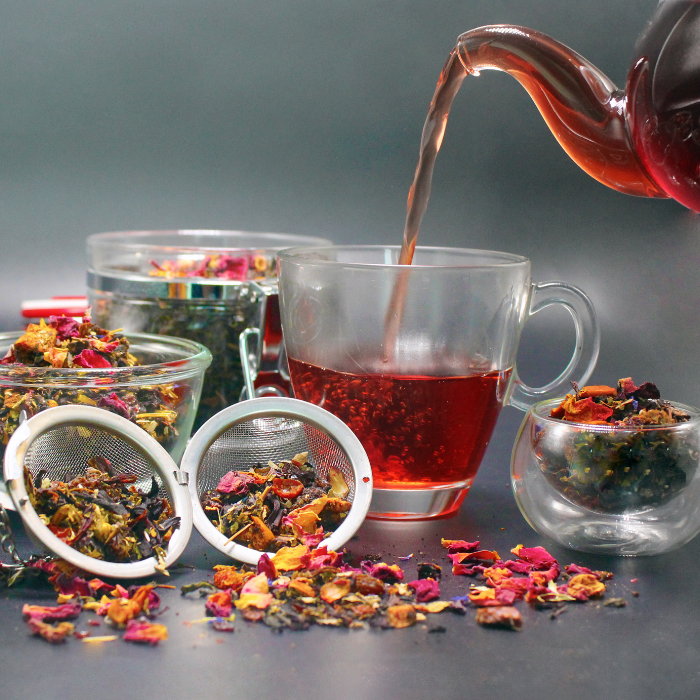Red herbal tea poured into a glass cup beside infusers of loose-leaf tea and dried flower petals