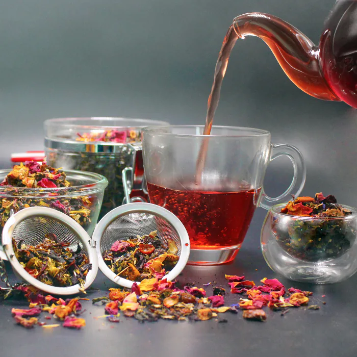 Red herbal tea poured into a glass cup beside infusers of loose-leaf tea and dried flower petals
