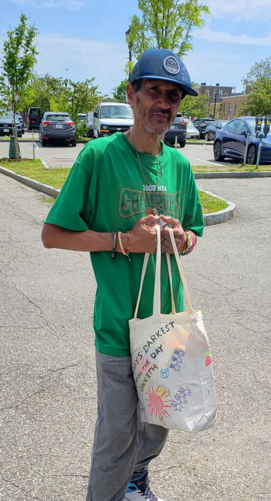 Man in a green T-shirt and cap holding a hand-decorated canvas tote bag in a sunny parking lot