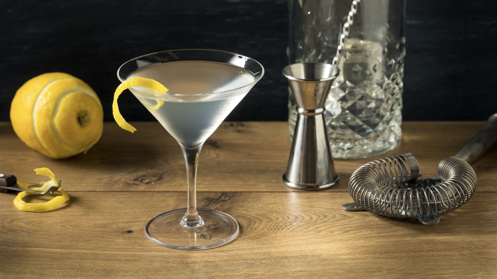 Martini with a lemon twist on a wooden bar beside a jigger, mixing glass and strainer