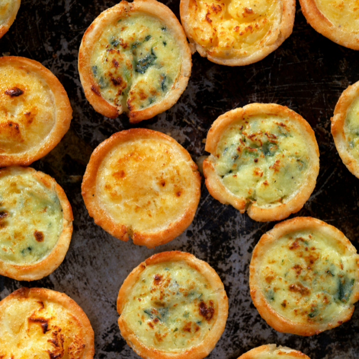 Mini spinach and cheese quiches with golden flaky crusts on a dark baking tray
