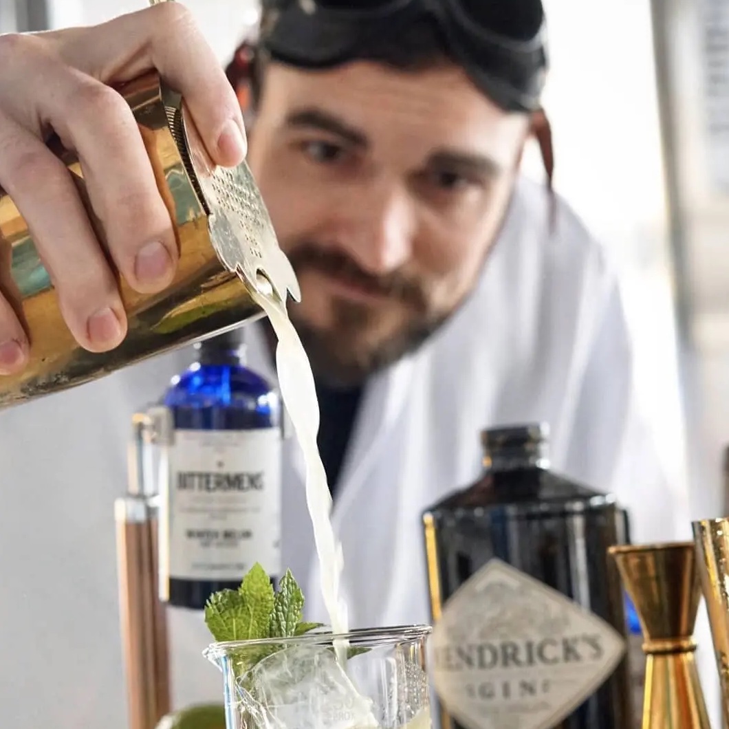 Mixologist T.J. Fink pouring a spirit at a home bar stocked with bottles including Hendrick's gin