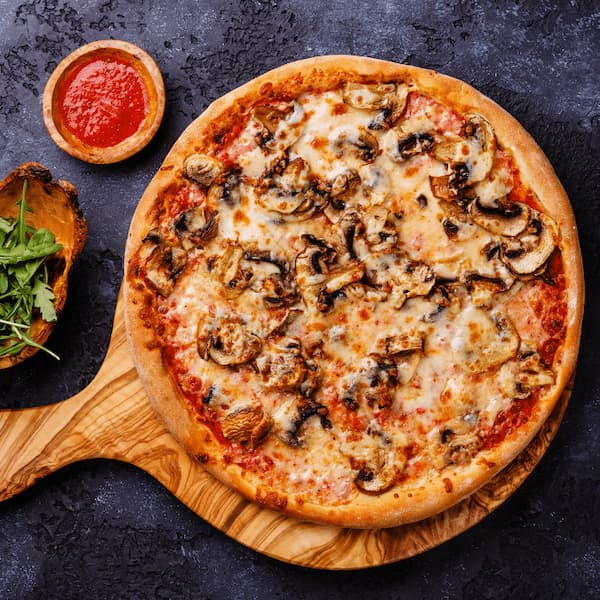 Mushroom and mozzarella pizza on a wooden peel with a bowl of tomato sauce and arugula