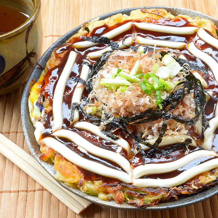 Okonomiyaki savory pancake with brown sauce, mayo drizzle, bonito flakes, nori and scallions on a bamboo mat