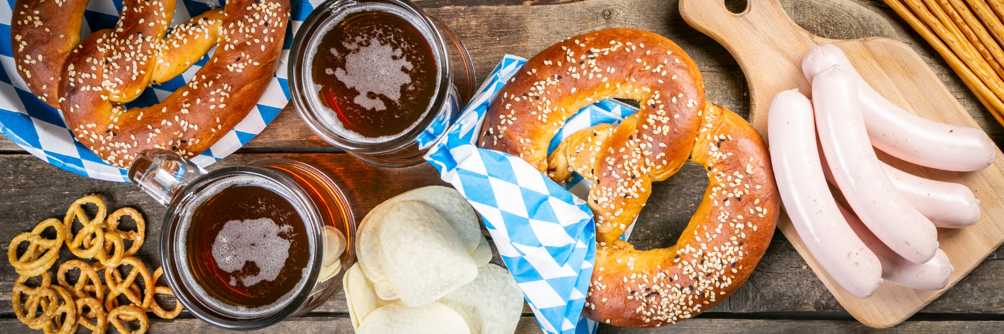 Oktoberfest spread of soft pretzels, beer mugs, white sausages and chips on blue checked napkins