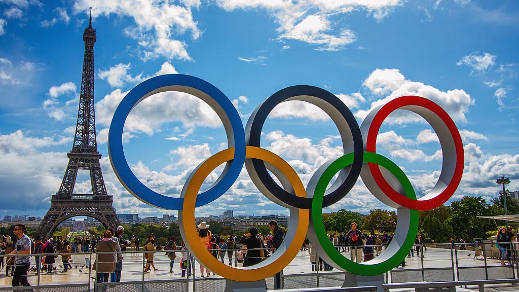 Olympic rings sculpture on a Paris plaza with the Eiffel Tower behind and visitors walking past