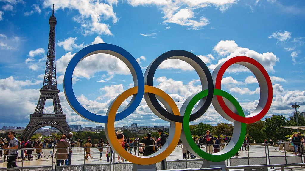 Olympic rings sculpture on a Paris plaza with the Eiffel Tower behind and visitors walking past