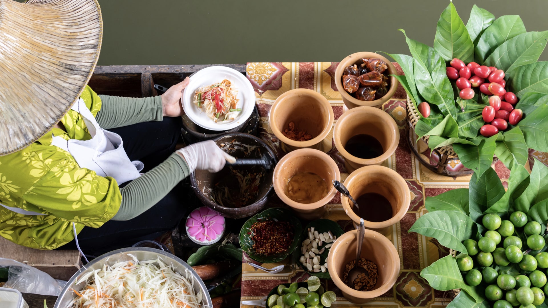 Overhead view of a cook in a conical hat plating food among clay pots of sauces, chilies and fresh herbs