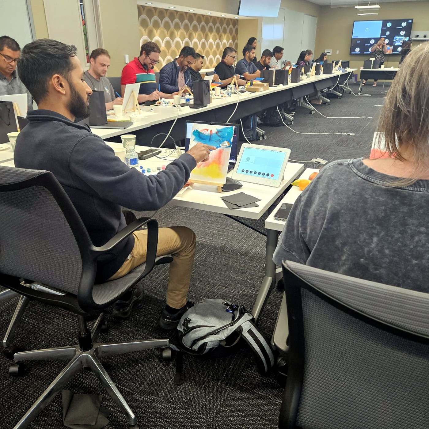 Coworkers painting on canvases at a long office table lined with laptops and paint palettes