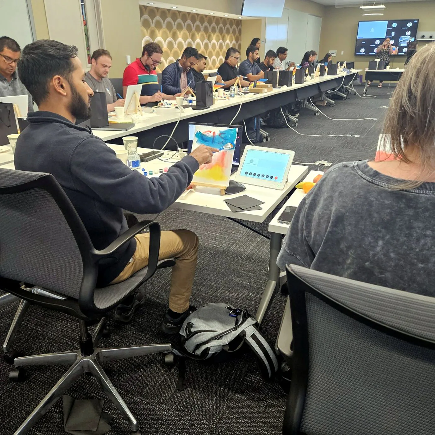Coworkers painting on canvases at a long office table lined with laptops and paint palettes