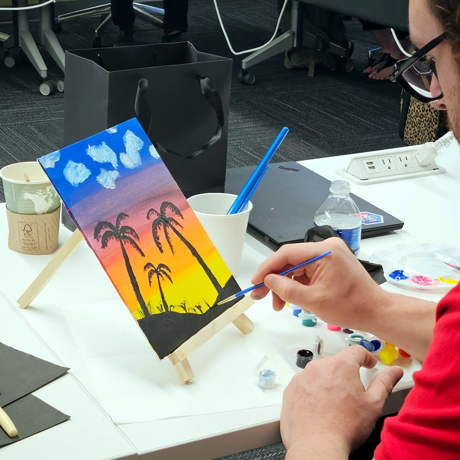 Hands painting palm tree silhouettes onto a sunset canvas propped on a small wooden easel