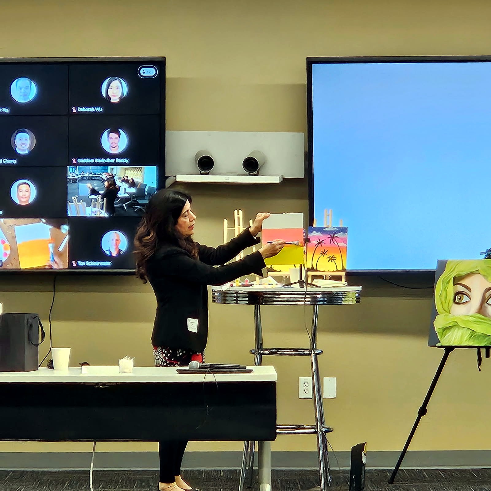 Host leading a paint session beside a wall screen of remote participants and finished canvases on easels