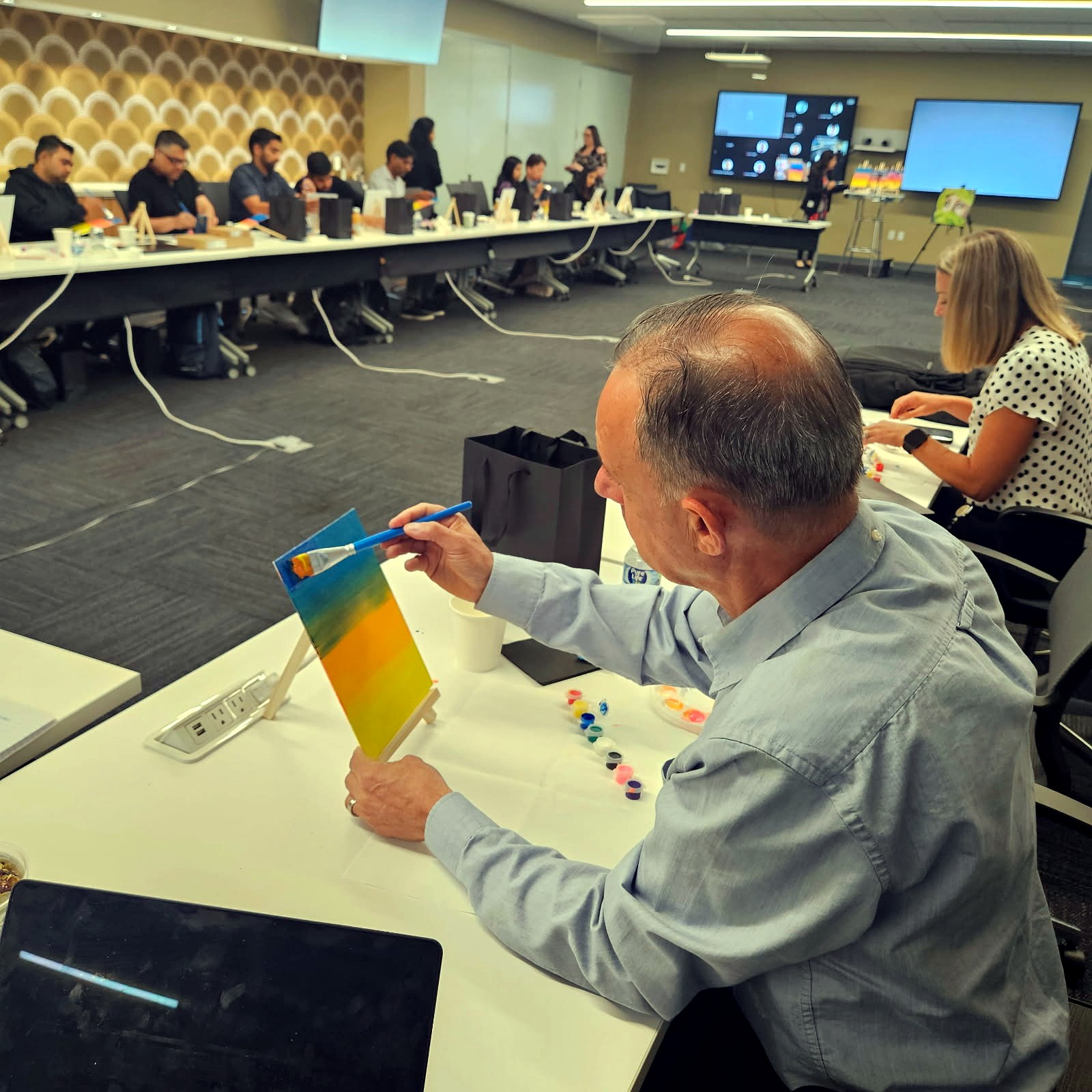 Man holding up his rainbow gradient painting at a conference room table during a team paint session