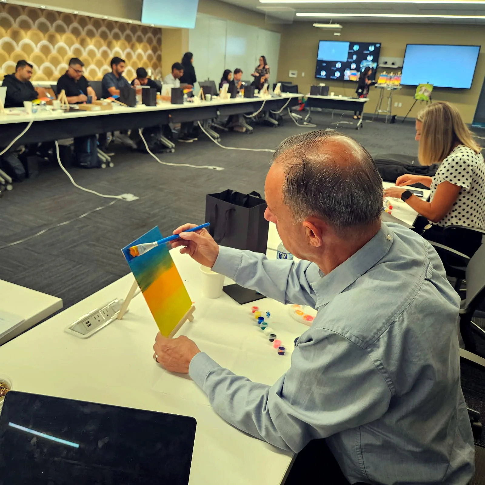 Man holding up his rainbow gradient painting at a conference room table during a team paint session