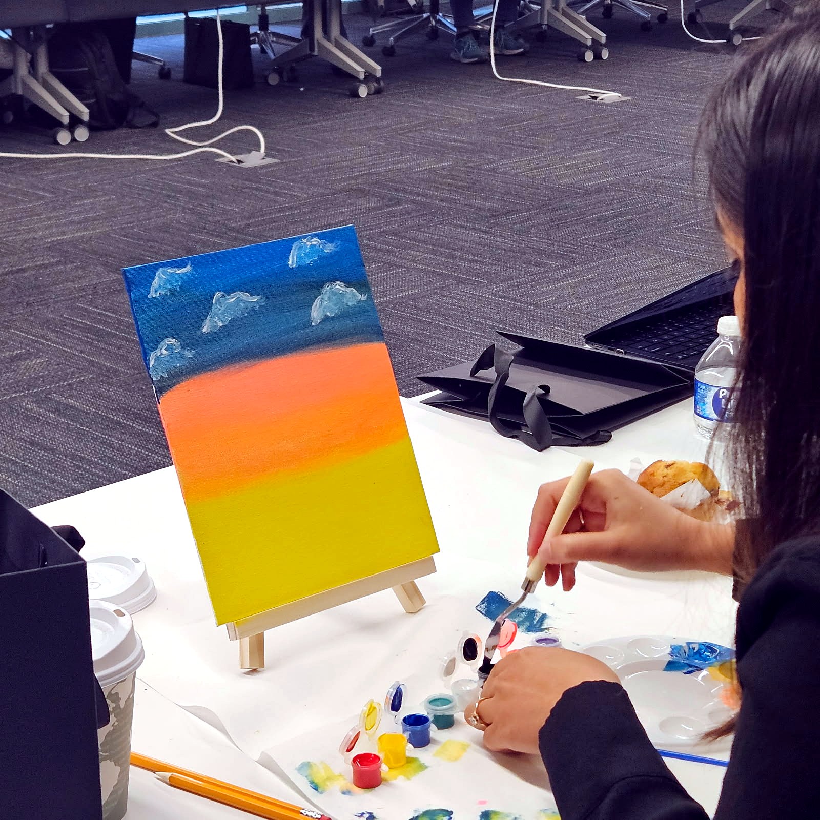 Overhead view of an orange sunset canvas on a tabletop easel beside dabs of paint on a palette