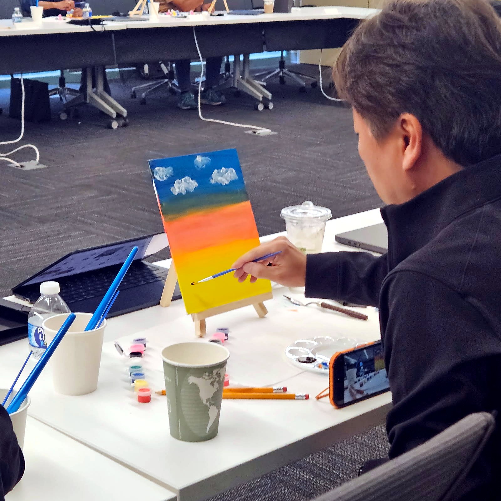 Over-the-shoulder view of someone painting an orange sunset canvas on a small tabletop easel