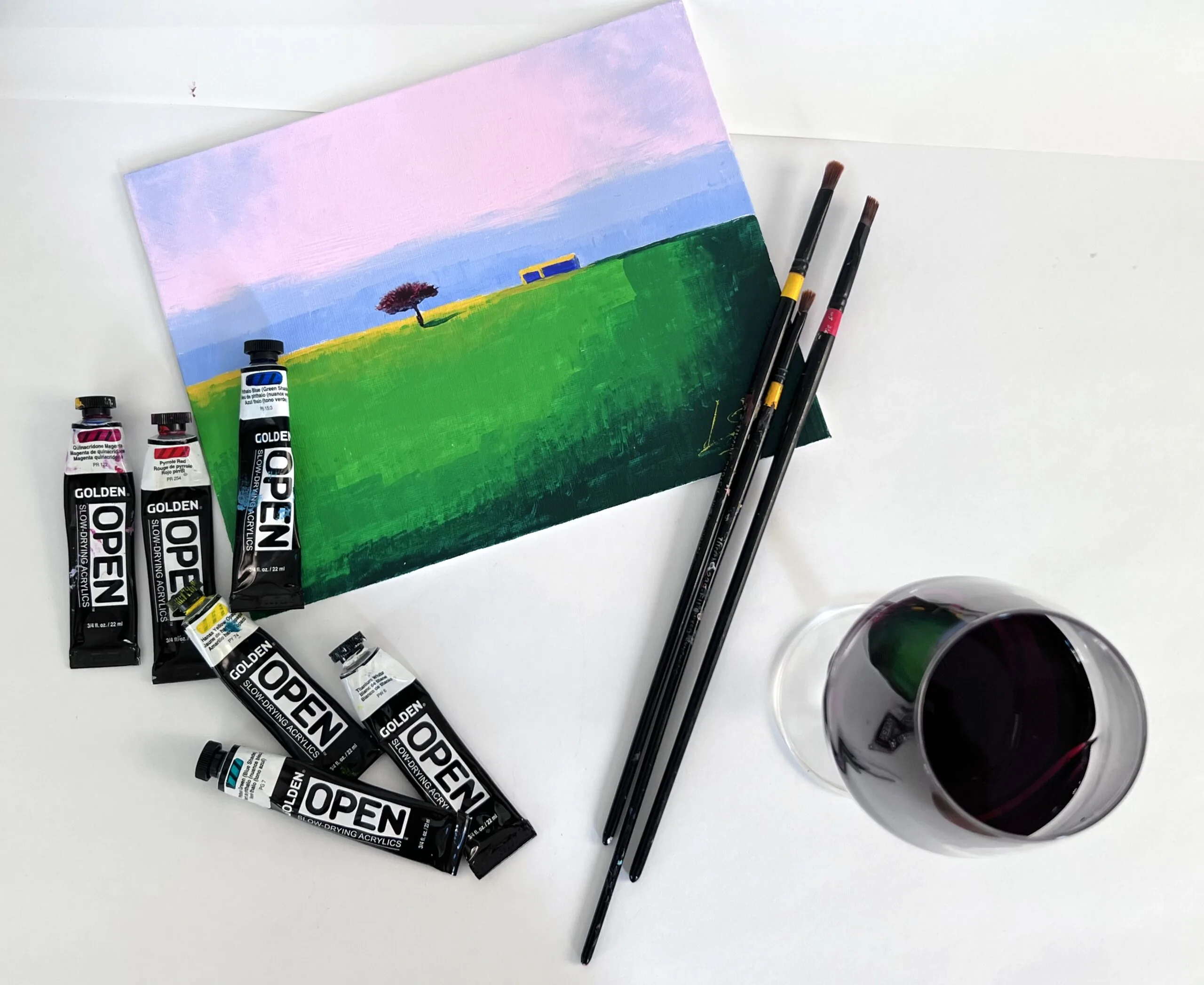 Finished landscape painting with acrylic paint tubes, brushes and a glass of red wine