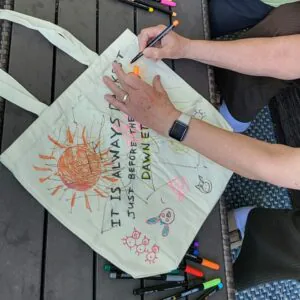Hands lettering a canvas tote bag with markers: It is always darkest just before the dawn