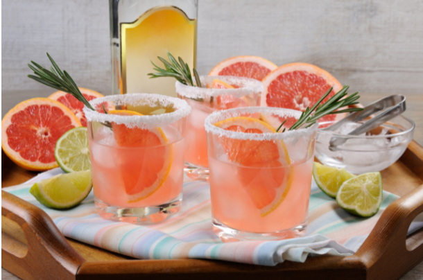 Pink grapefruit palomas in salt-rimmed glasses with rosemary, beside fresh grapefruit and lime