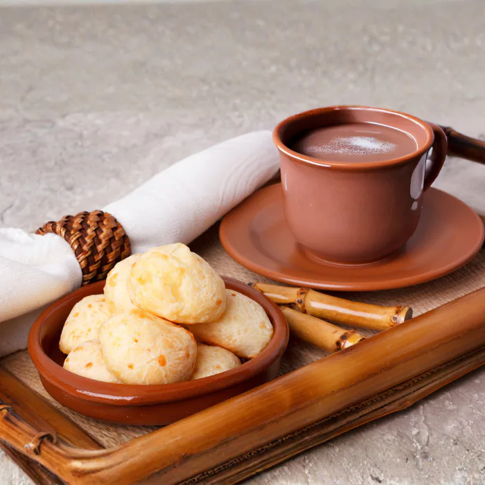 Brazilian cheese breads in a terracotta dish beside a cup of thick hot chocolate on a bamboo tray
