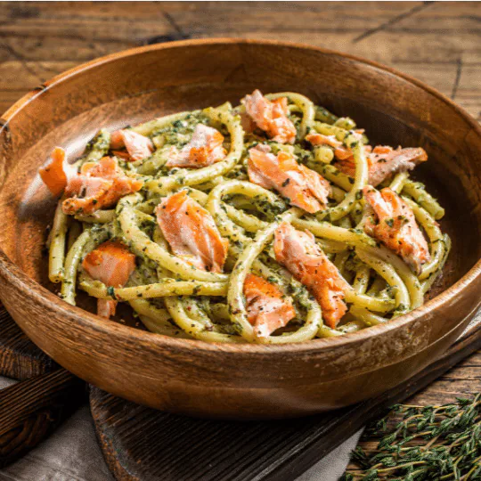Long pasta tossed with green pesto and flakes of salmon in a wooden bowl