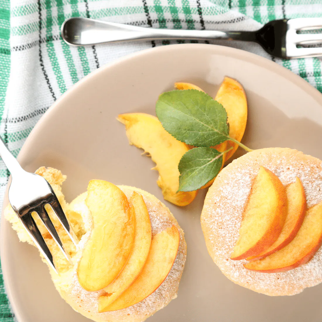 Small round tea cakes topped with fresh peach slices and powdered sugar, with a forkful lifted away