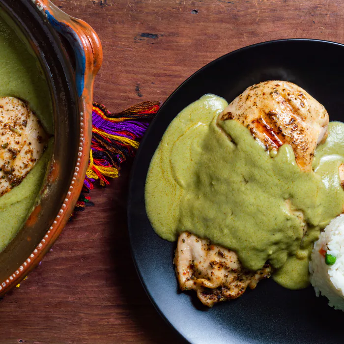 Seared chicken under green pumpkin seed pipián mole on a black plate with rice, beside a clay pot