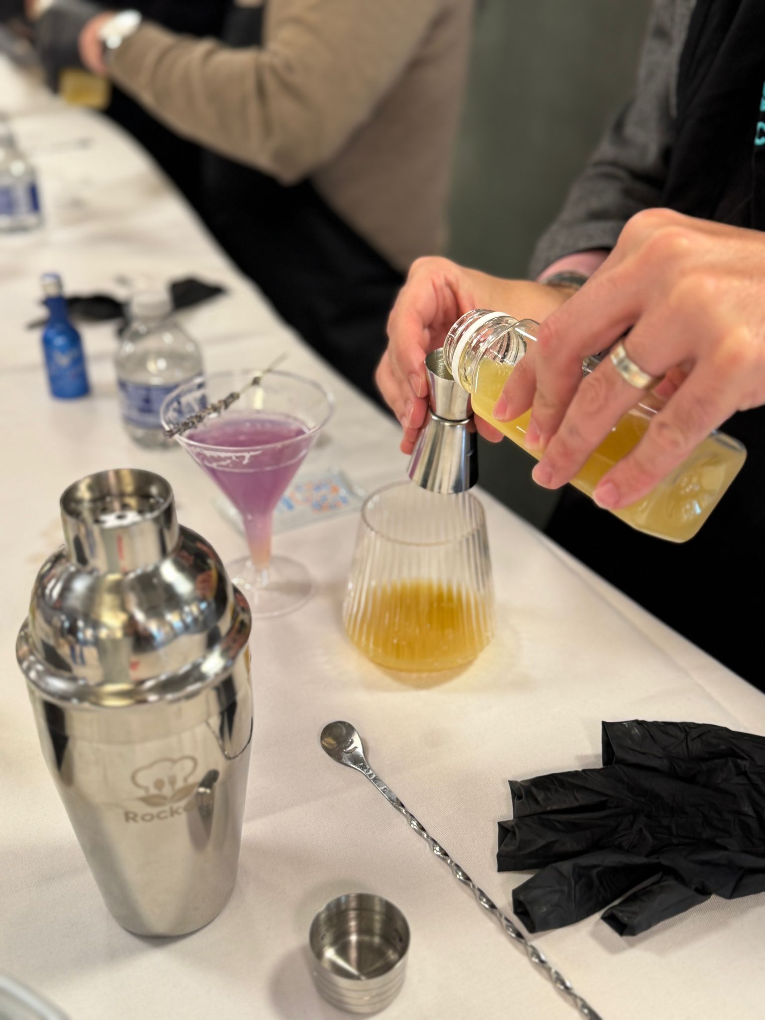 Hand pouring juice into a ribbed mixing glass beside a Rockoly cocktail shaker and a purple cocktail