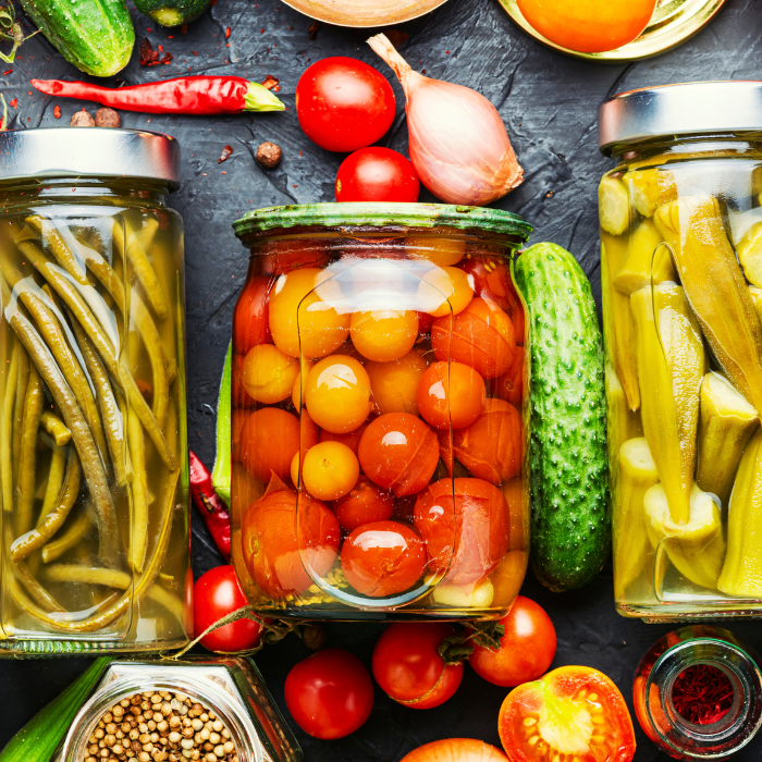 Jars of pickled cucumbers, beans and tomatoes surrounded by fresh vegetables, chiles and garlic