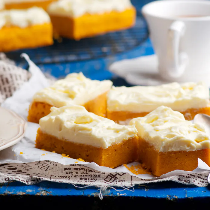 Squares of pumpkin bar under thick cream cheese frosting on parchment, with a cup of coffee behind