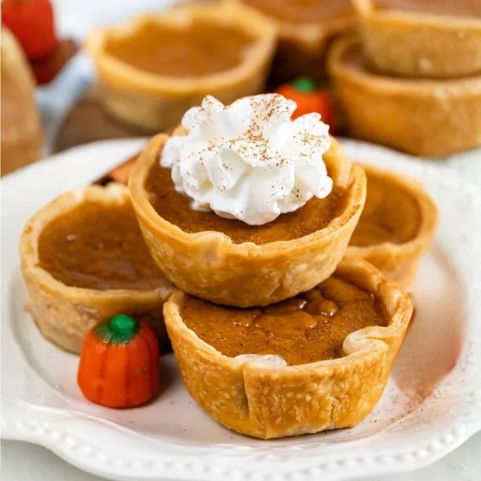 Mini pumpkin pies stacked on a white plate, the top one piped with whipped cream and dusted with cinnamon