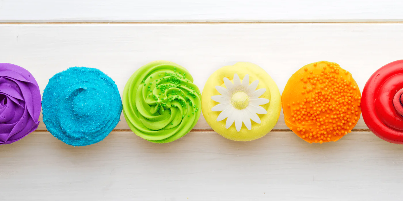 Row of cupcakes frosted in purple, blue, green, yellow, orange and red on white wood