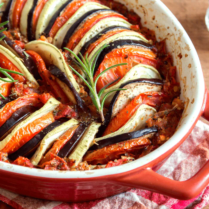 Ratatouille of thinly sliced eggplant, zucchini and tomato layered in a red baking dish with rosemary